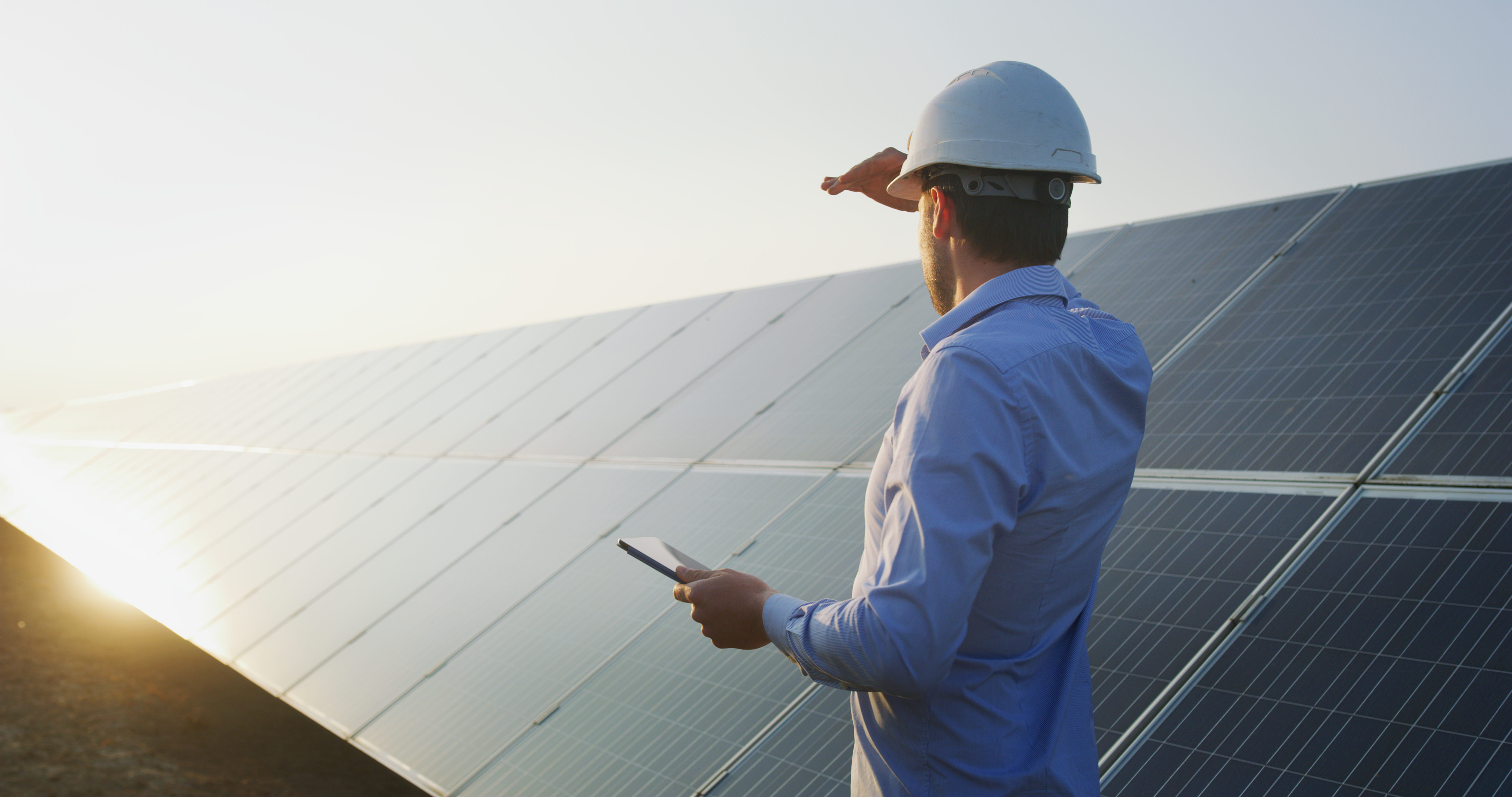 An engineer with a tablet and a hard hat, looking at a row of solar panels.